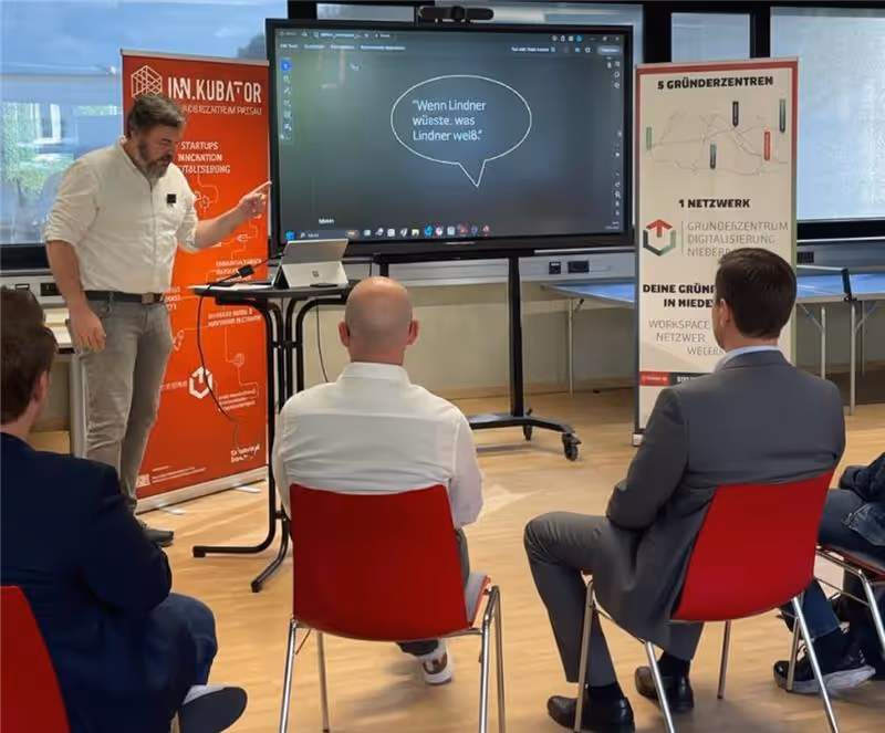 A man standing and presenting to an audience seated in red chairs with a large screen displaying a German quote in a speech bubble in a modern meeting room.